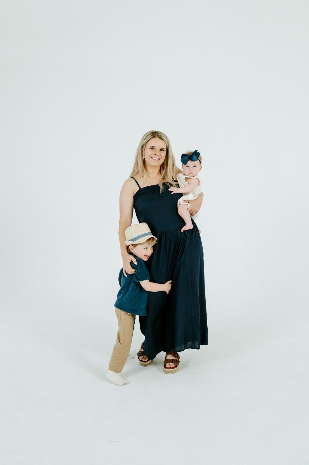 A smiling woman in a navy dress holds a baby girl while a young boy in a hat hugs her side in a studio.
