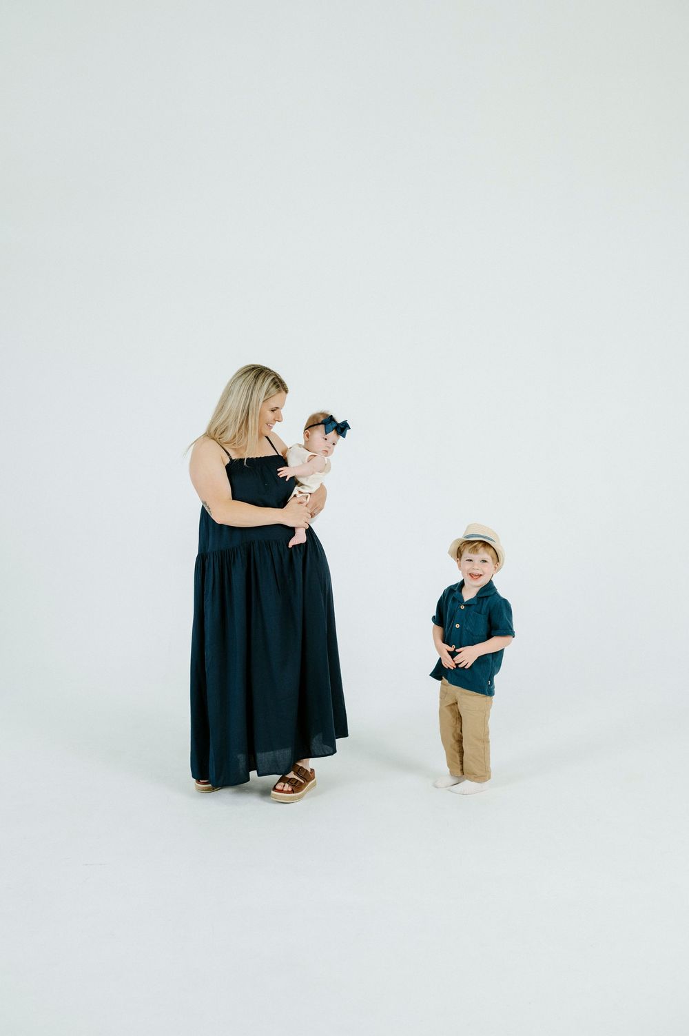 A mother in a navy dress holds her baby girl while her young son in a straw hat stands beside her on white background.