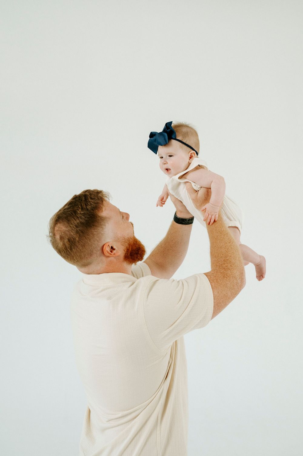 A red-haired father lifts his baby girl wearing a navy bow headband against a white background.