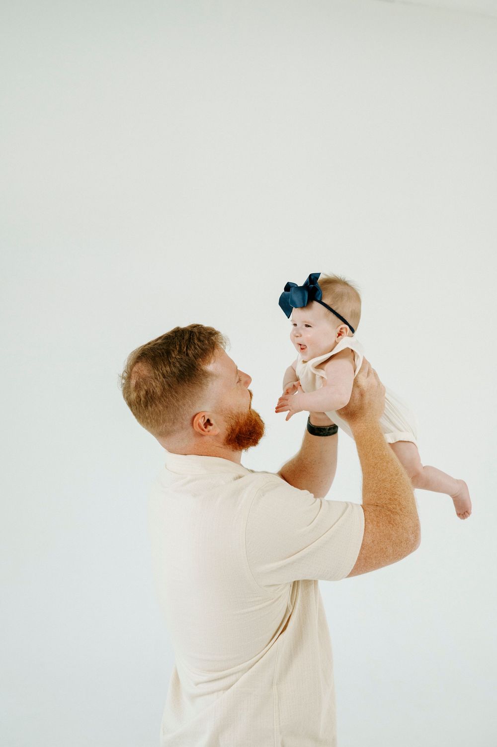 Red-haired bearded father lifting smiling baby girl with navy bow headband against white background.