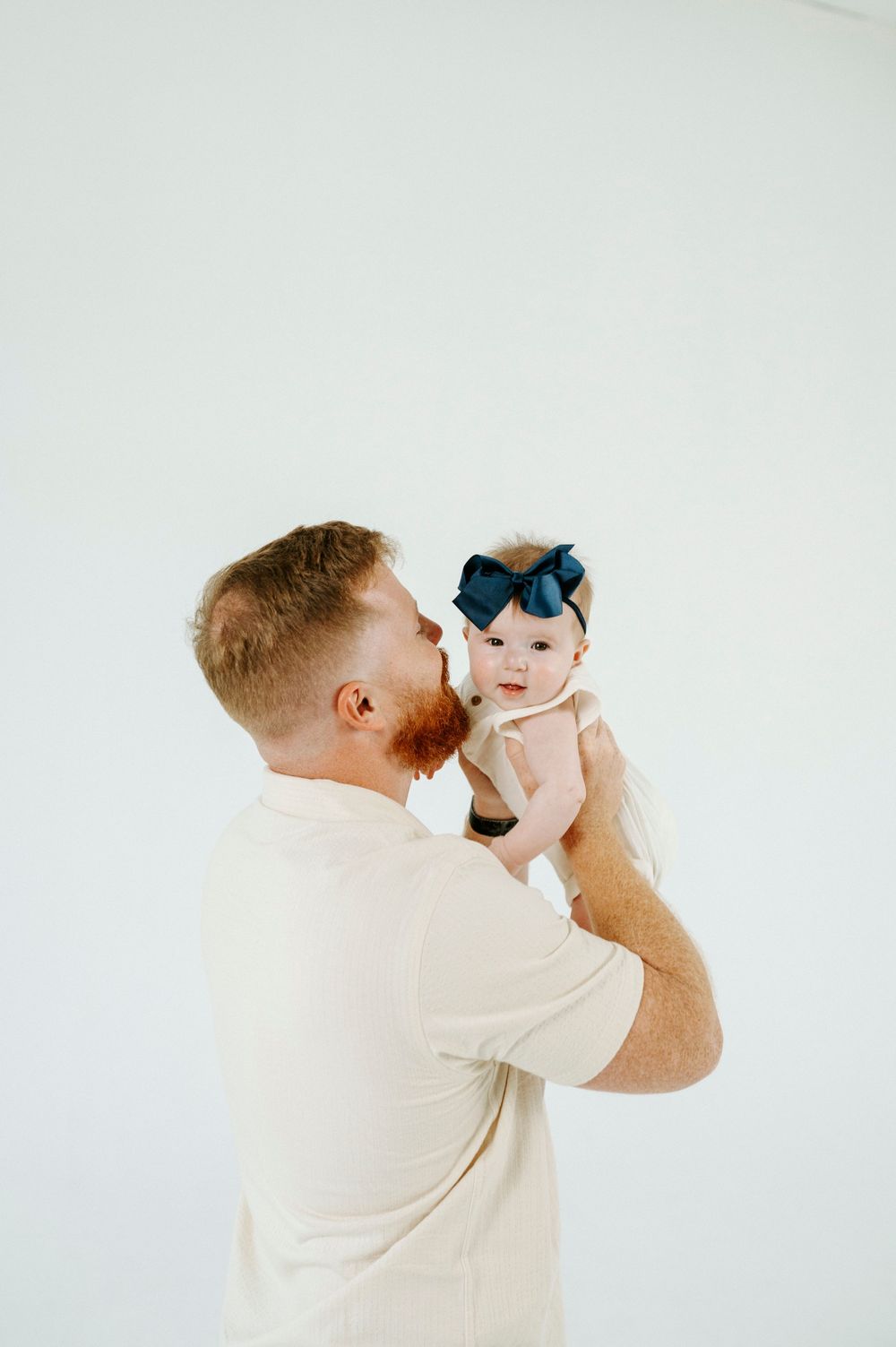 Red-haired father holding up his baby girl wearing a navy bow headband against a white background.