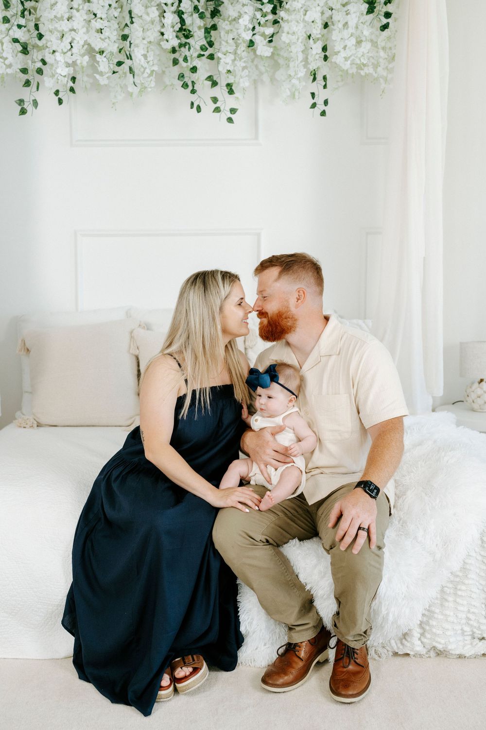 A couple shares a tender moment while holding their newborn baby, seated on a white bed with floral decor.