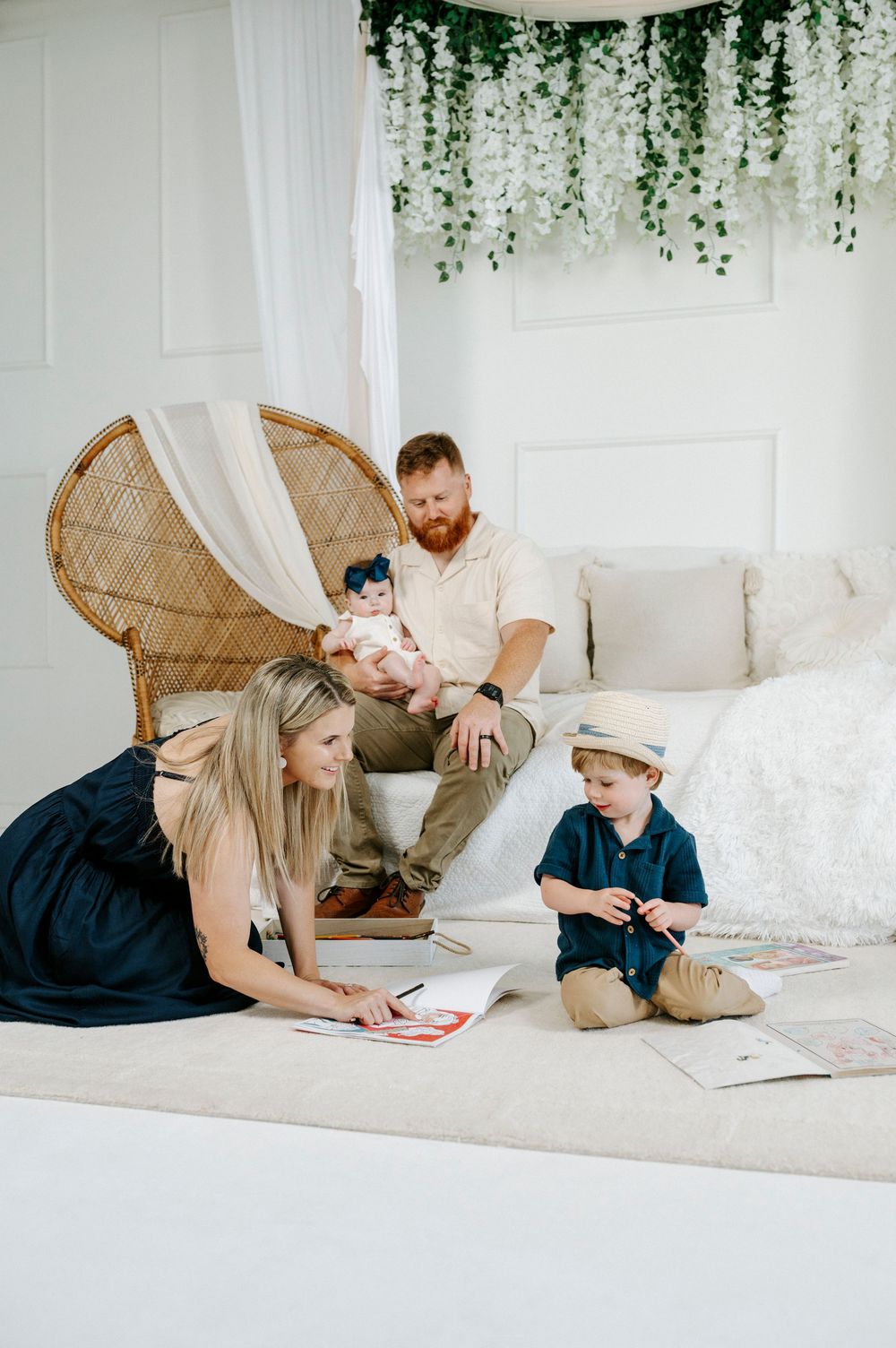 Family of four enjoying a cozy white room, mom and toddler drawing on floor while dad holds baby near wicker chair.