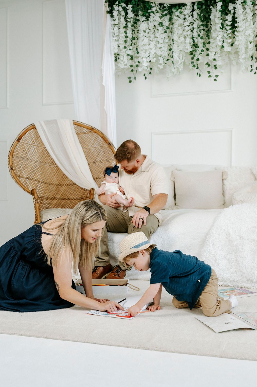 Family of four enjoying a cozy white bedroom, parents with newborn and toddler boy drawing on floor.