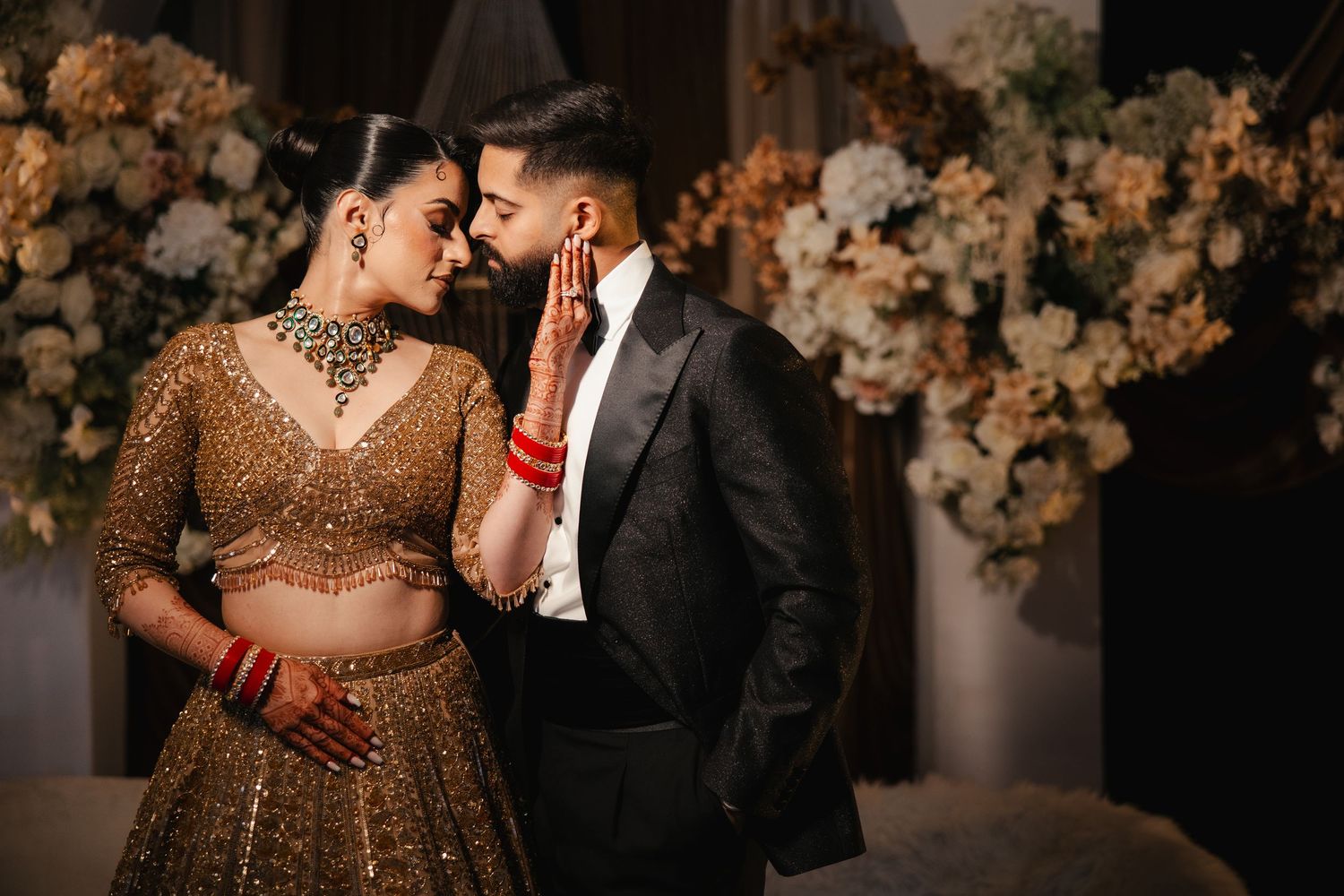Bride in gold lehenga and groom in black suit share romantic moment at Indian wedding ceremony with floral backdrop.
