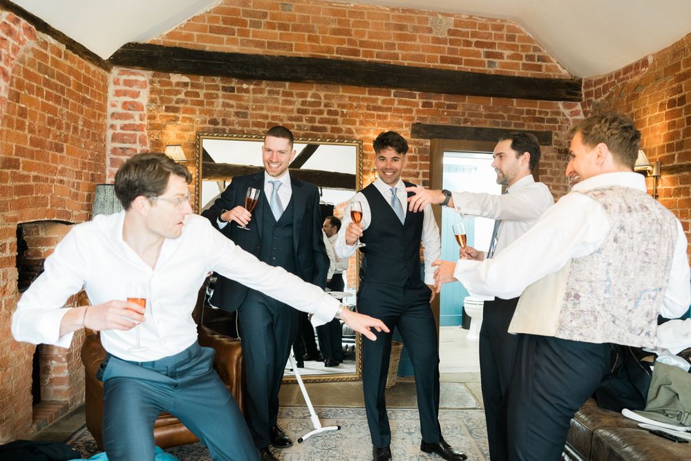 Group of groomsmen celebrating together in a rustic brick-walled room before a wedding ceremony.