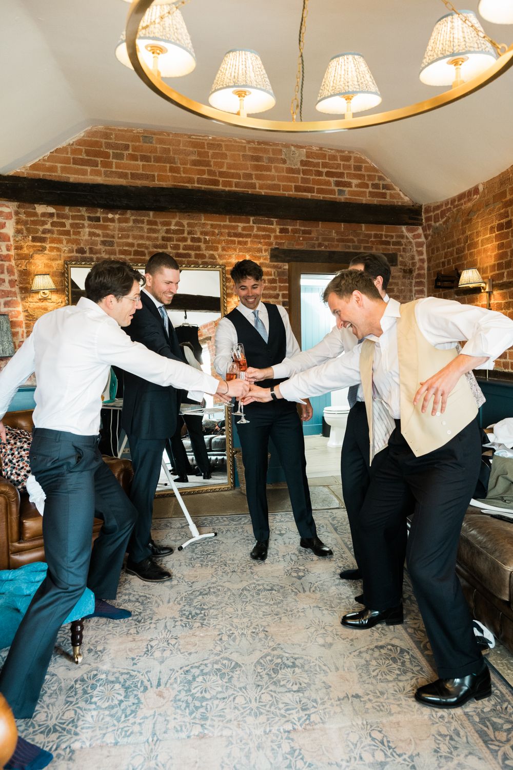 Groomsmen in suits toasting with champagne glasses in a rustic brick-walled room before a wedding ceremony.