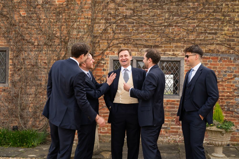 Five groomsmen in navy suits chat outdoors beside a rustic brick wall at a wedding celebration.