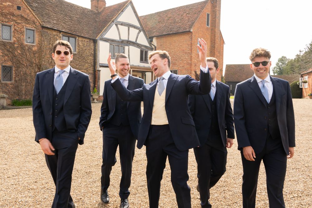 Four groomsmen in navy suits and light blue ties walk together outside a Tudor-style venue on a sunny day.