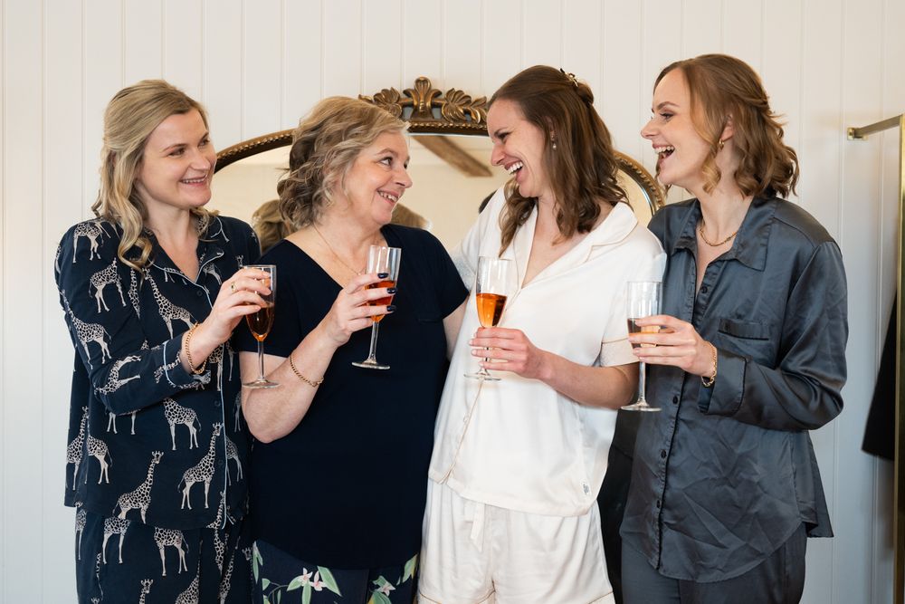 Four women in pajamas laughing and toasting with champagne glasses during a bridal party getting ready celebration.