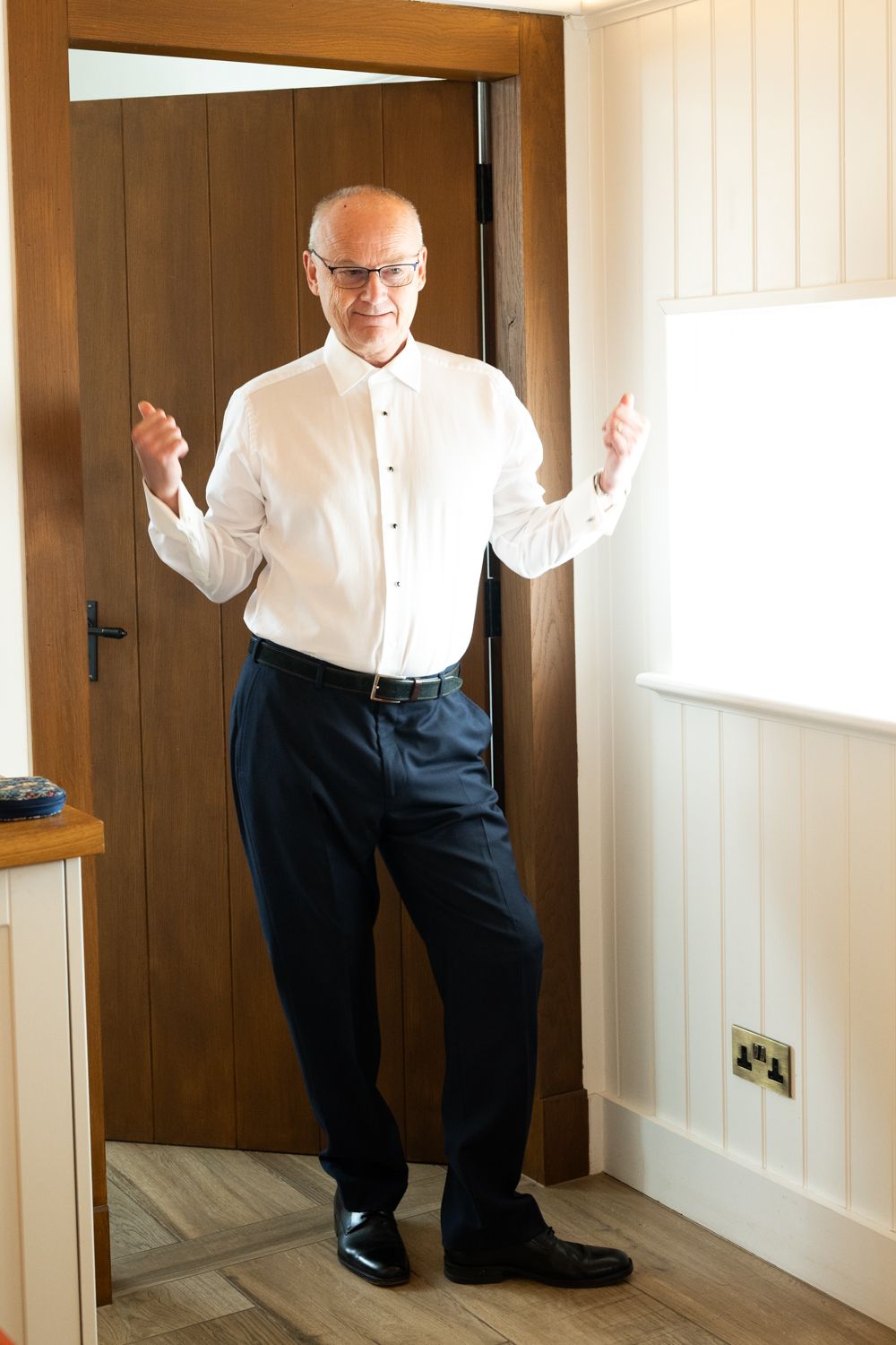 Older man in white dress shirt and dark trousers posing confidently in a bright doorway.