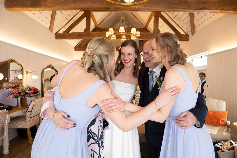 Bride in white gown shares a joyful group hug with bridesmaids in blue dresses and a guest at a rustic wedding venue.