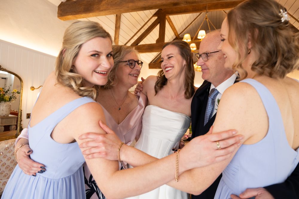 Bride in white gown shares joyful group hug with bridesmaids in blue dresses and family members at wedding.