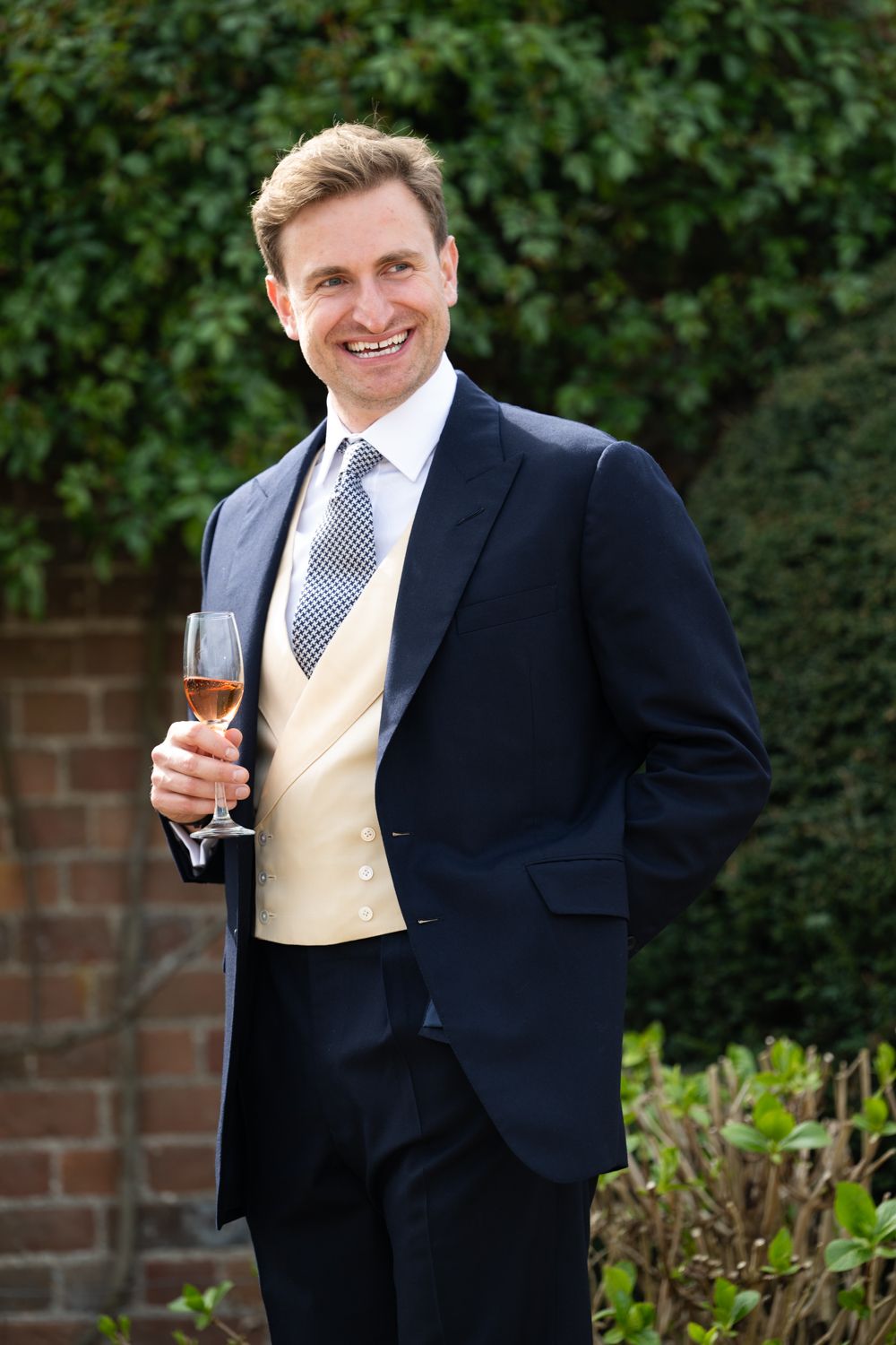 Smiling man in navy suit with cream waistcoat holding a glass of rose wine outdoors at a formal event.