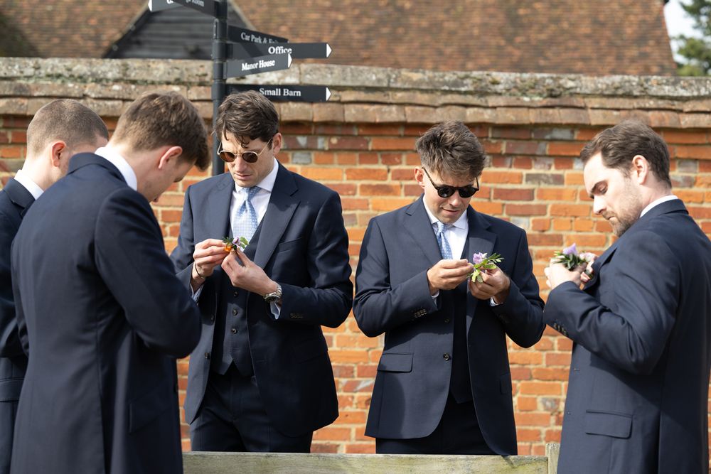 Group of young men in navy suits pinning boutonnieres outside a brick building at a wedding.