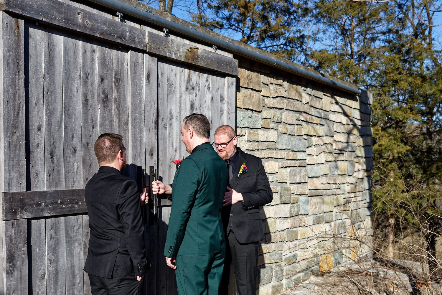 Three men in formal attire converse outside a rustic stone and wood barn on a sunny day.