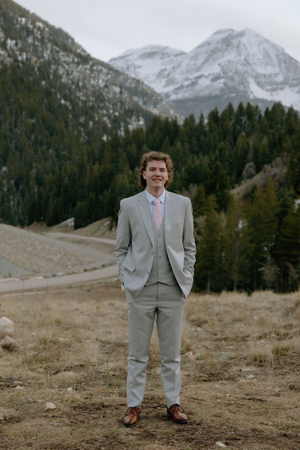 Smiling groom in gray suit with pink tie standing before snow-capped mountains and pine forest.