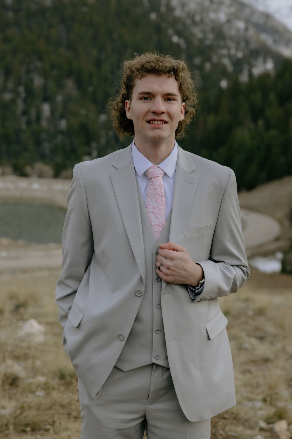 Young groom in gray three-piece suit with pink floral tie poses outdoors with mountain backdrop.