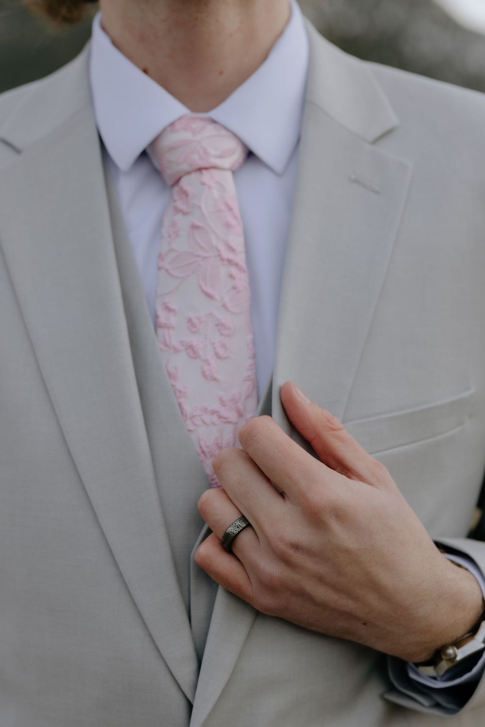 Groom in light gray suit adjusting pink floral tie, wearing wedding ring, close-up detail shot.