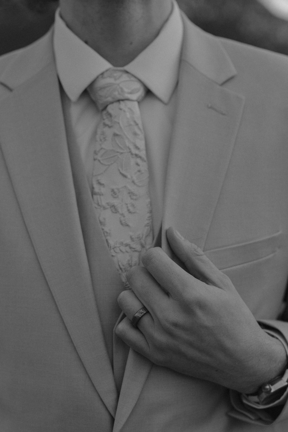 Black and white close-up of groom adjusting floral tie, wedding ring visible on hand.