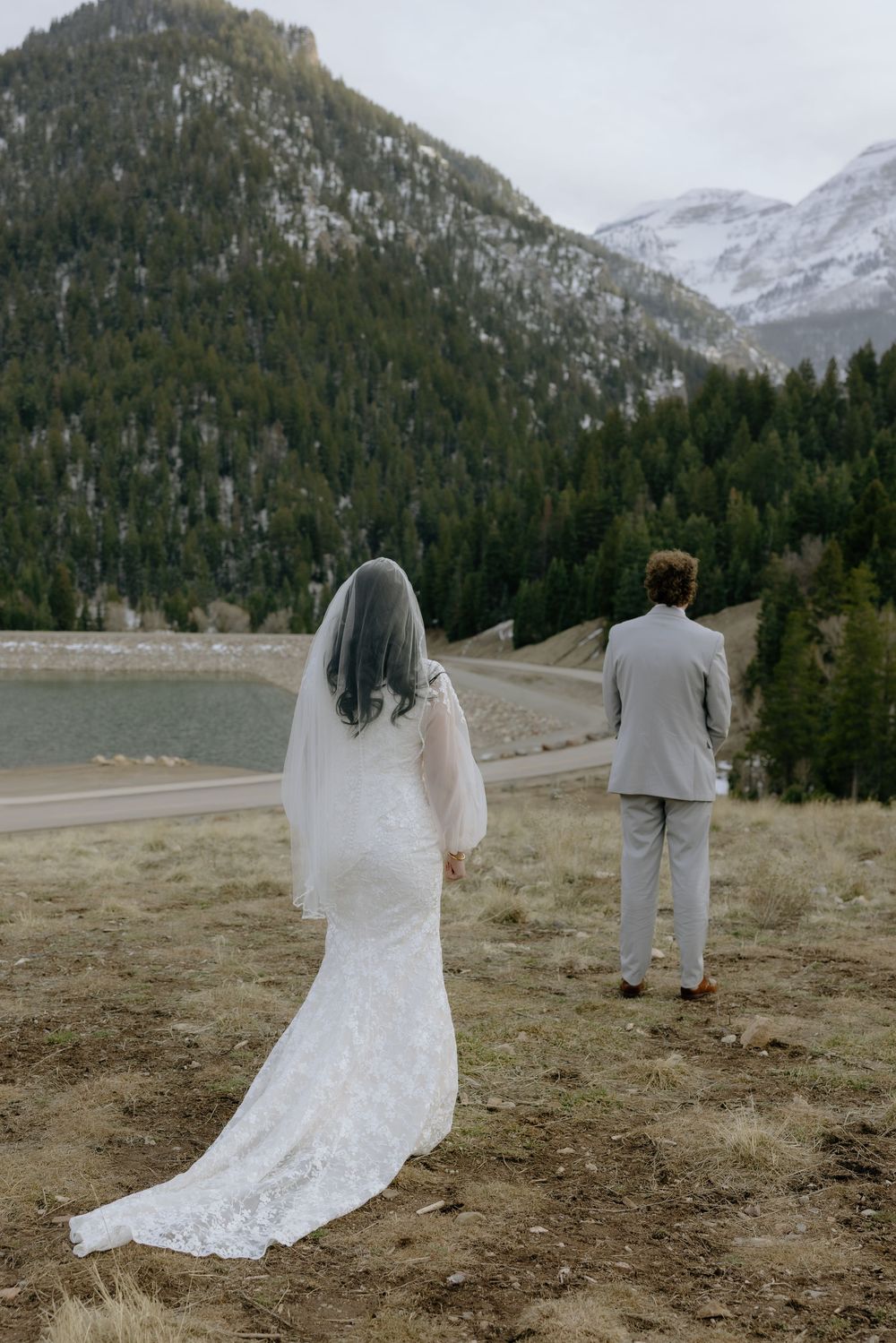 Bride and groom standing apart facing a mountain lake with snow-capped peaks and pine trees.