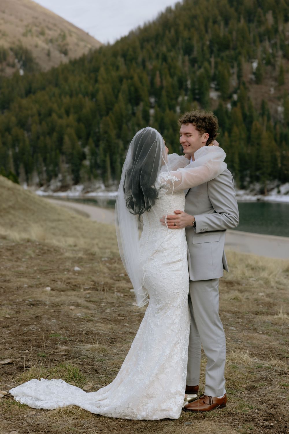 Bride in lace gown and veil embraces smiling groom in gray suit near mountain lake with pine forest backdrop.