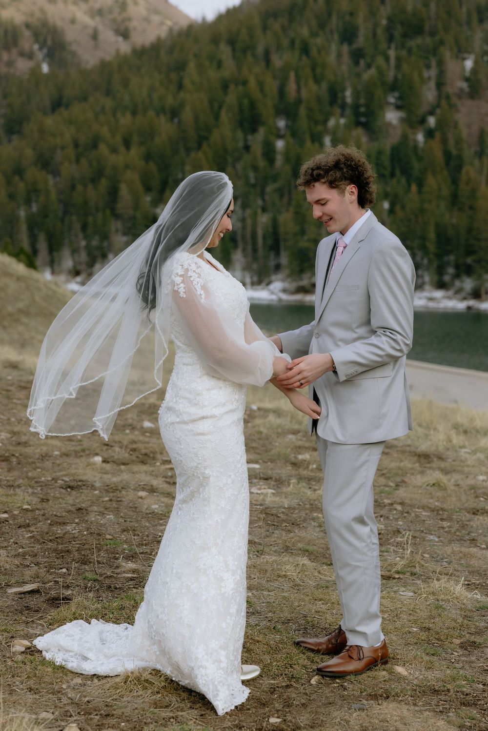 Bride and groom holding hands during outdoor wedding ceremony near mountain lake, bride in lace gown with veil.
