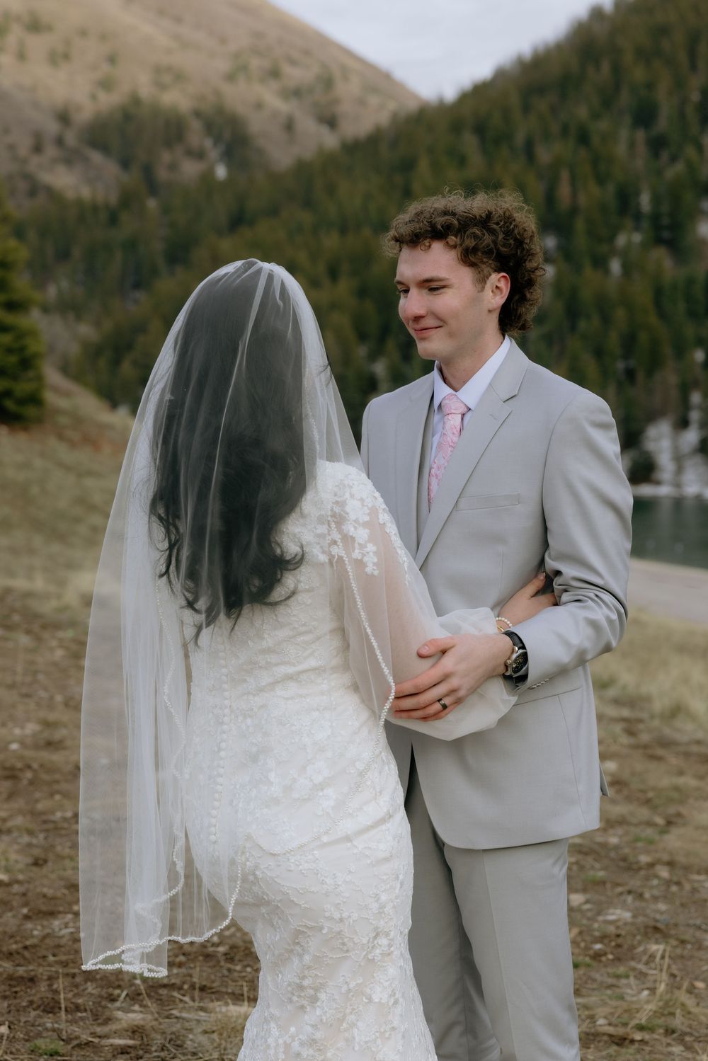 Groom in gray suit smiling at veiled bride in lace dress during outdoor mountain wedding.