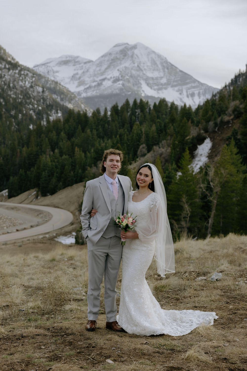 Bride and groom pose in wedding attire before snow-capped mountains and pine forest backdrop.