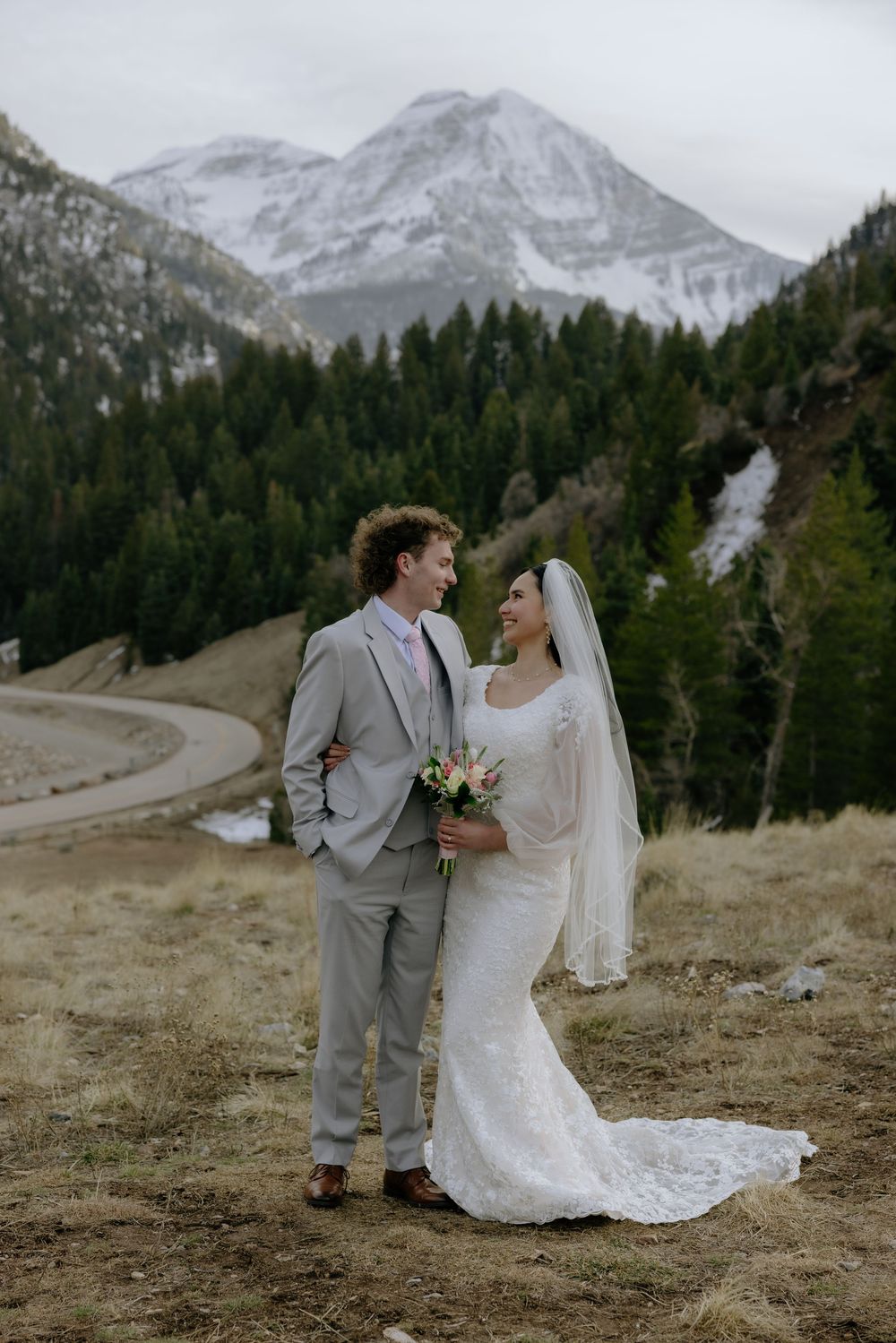 Bride and groom stand together smiling in mountain landscape with snow-capped peaks and pine forest backdrop.