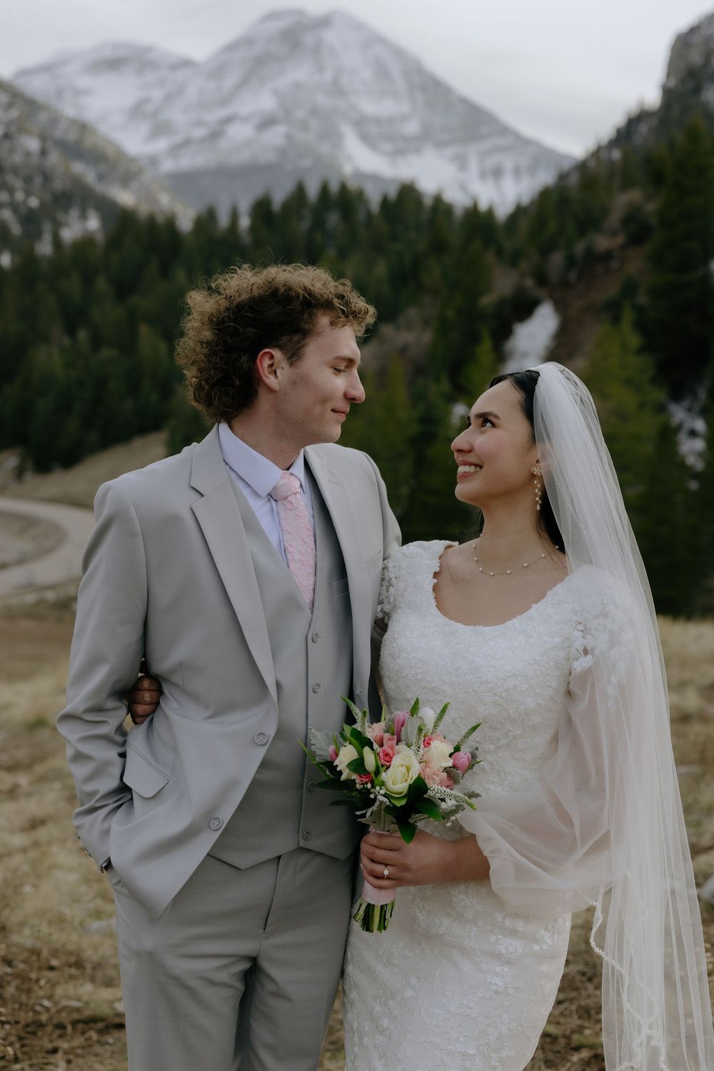 A bride and groom pose together in a mountain landscape, she holds a pink bouquet wearing a white gown with veil.