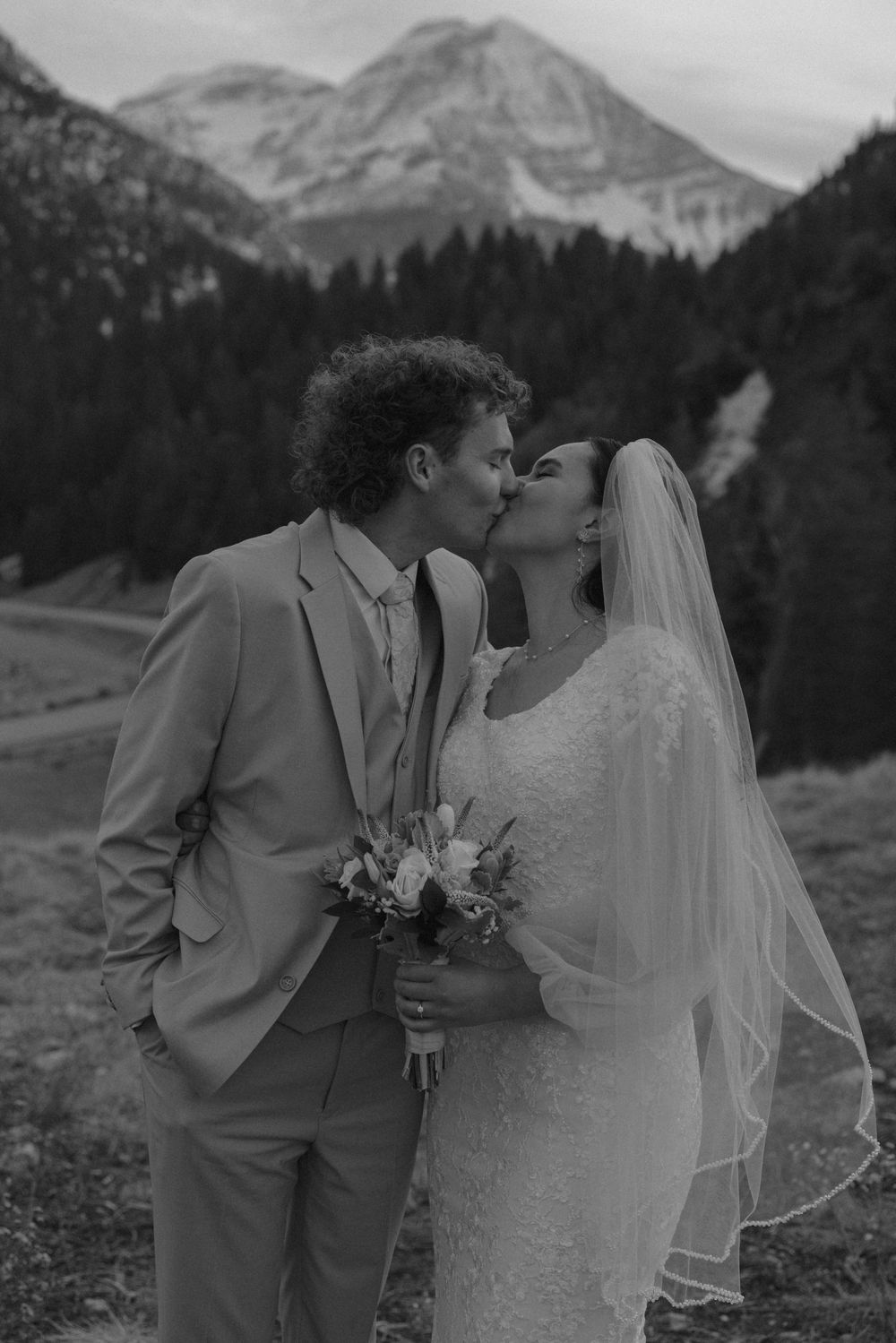 Black and white photo of a bride and groom kissing with mountain and forest backdrop on their wedding day.