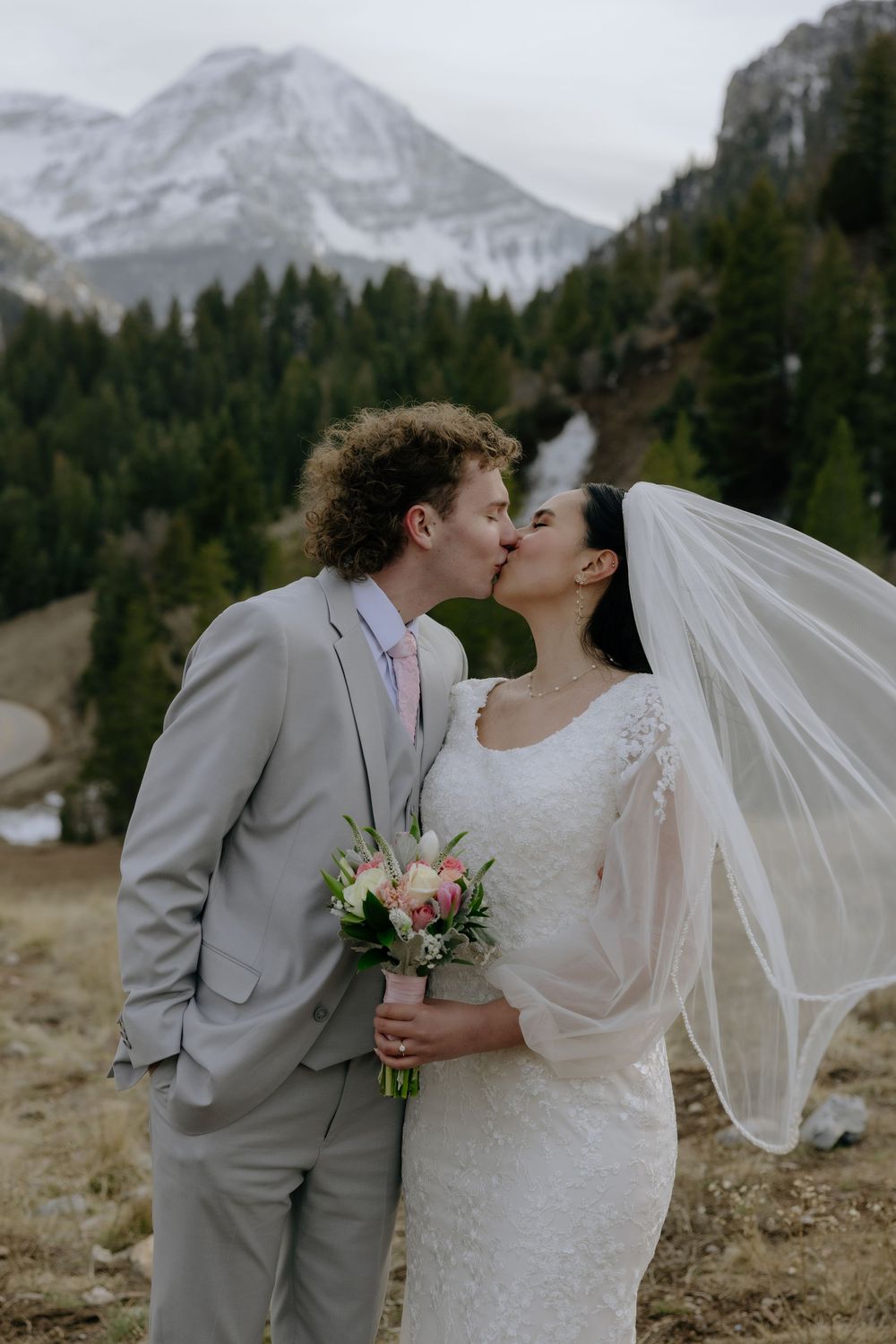 Bride and groom share a romantic kiss outdoors with stunning snowy mountain and evergreen forest behind them.
