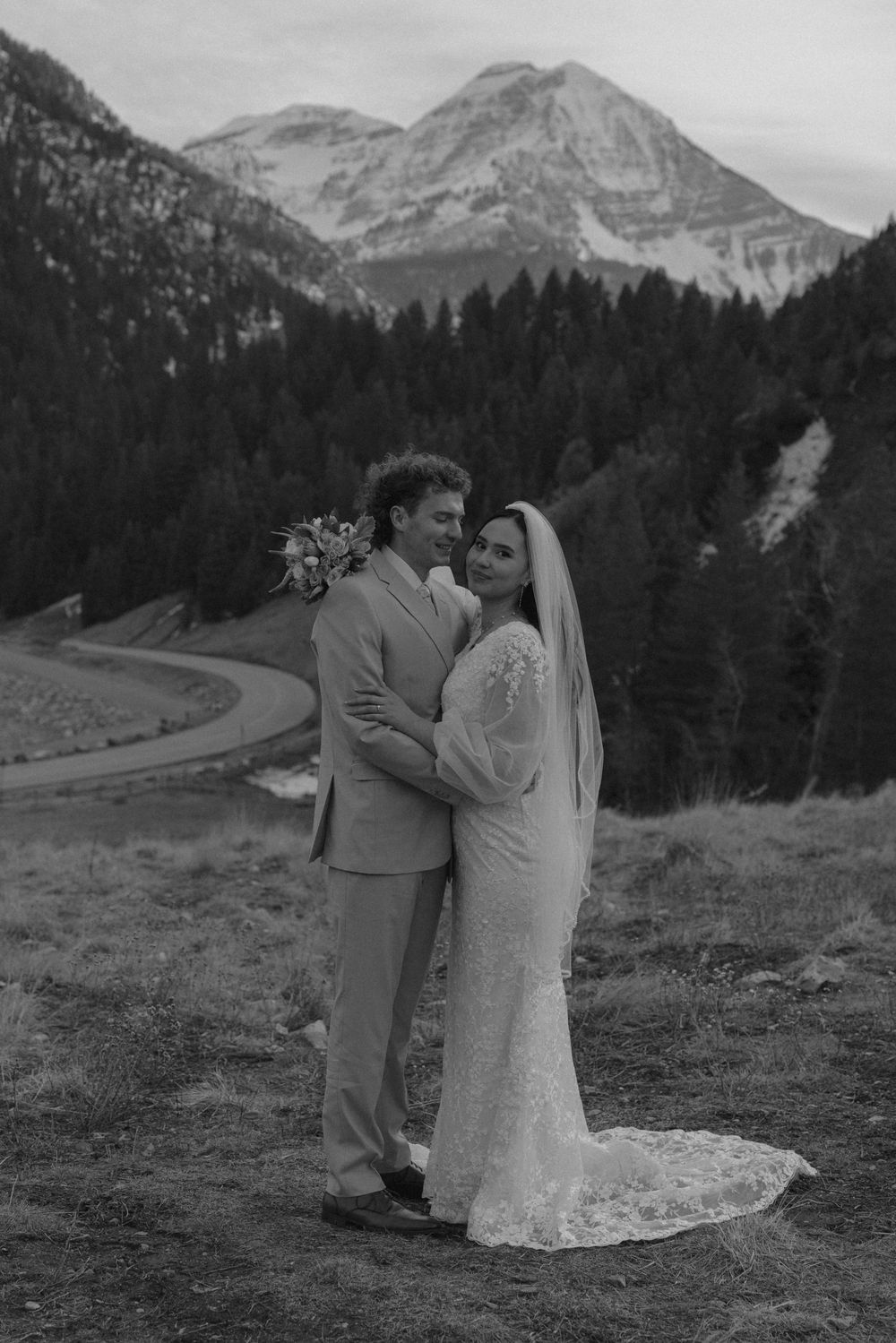 Black and white photo of a wedding couple embracing with mountain and forest scenery behind them.