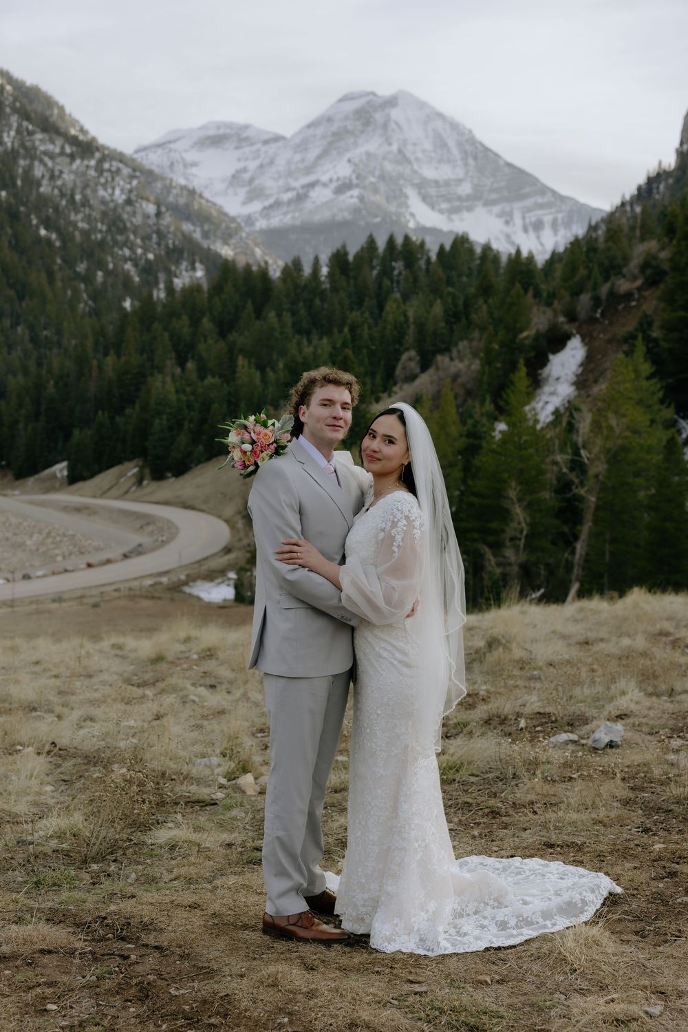 Bride and groom pose together in a mountain landscape with snow-capped peaks and evergreen forest backdrop.
