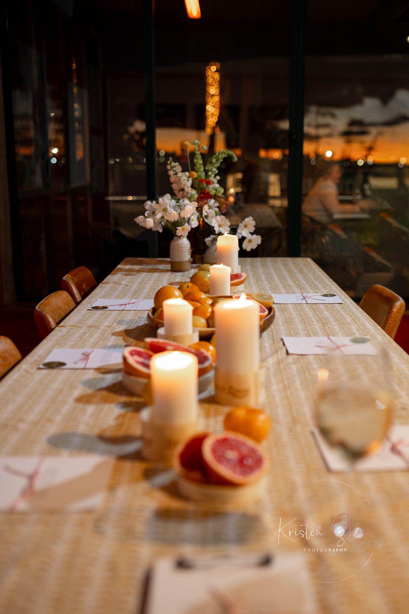 Elegant candlelit dinner table with white pillar candles, fresh grapefruit, oranges, and floral centerpiece at dusk.