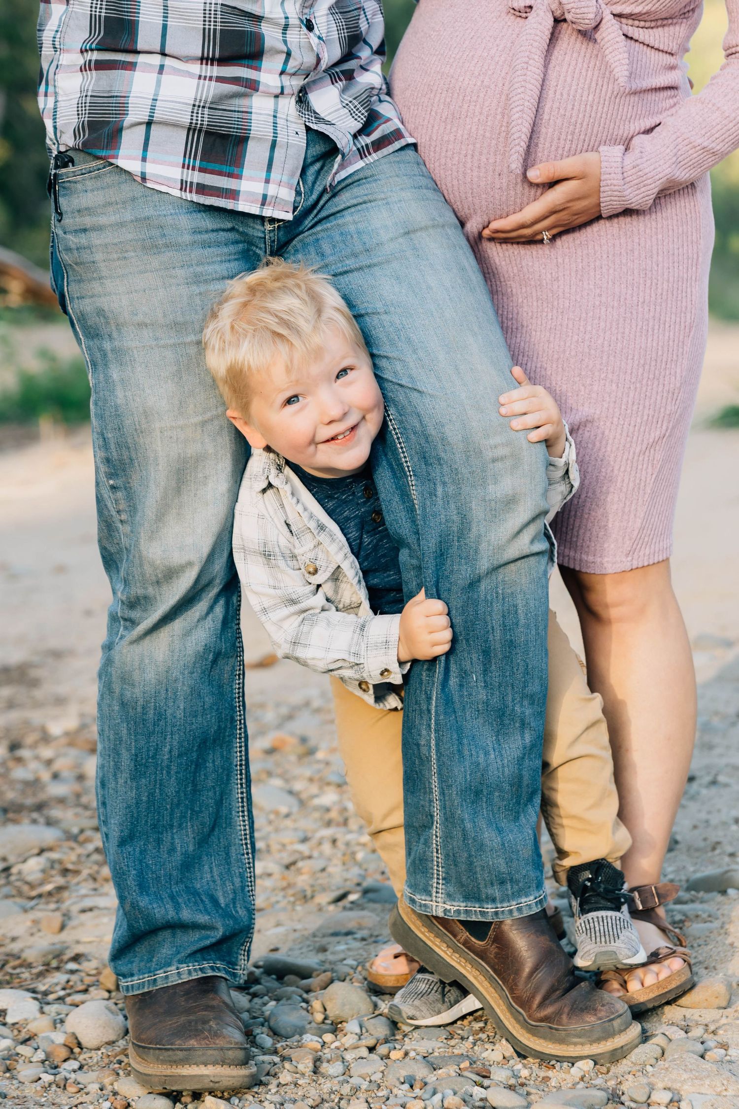 A young boy smiles while peeking between his parents legs outdoors, with his pregnant mother in a pink dress beside him.