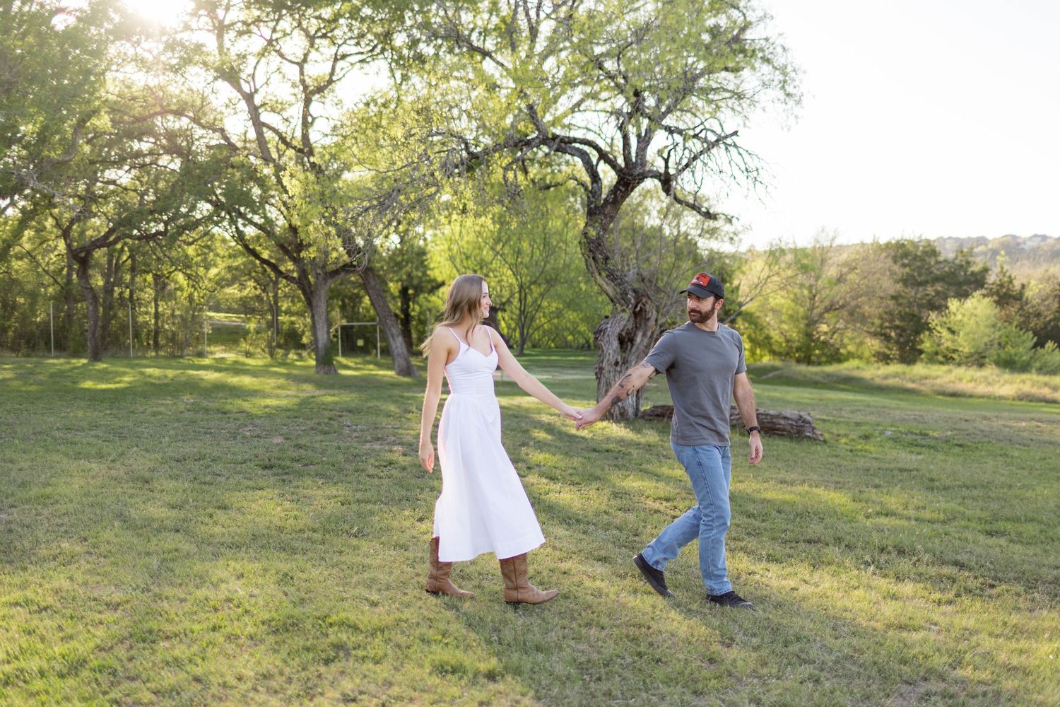 Couple holding hands walking through a sunny green meadow with trees, woman in white dress and boots, man in jeans captured by Nicole Parker