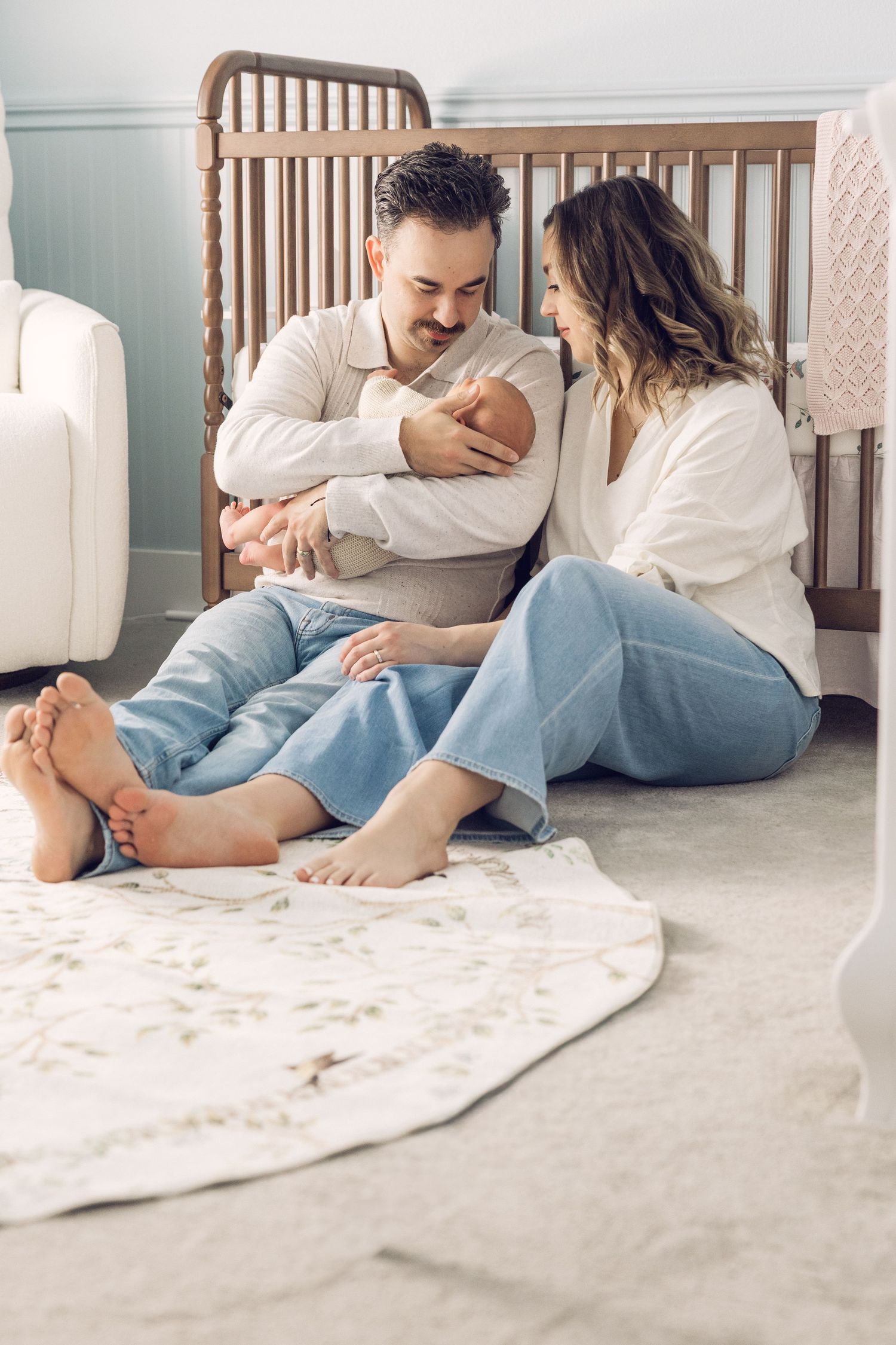 A couple sits barefoot on a nursery floor, smiling at their newborn baby held by the father near a wooden crib.