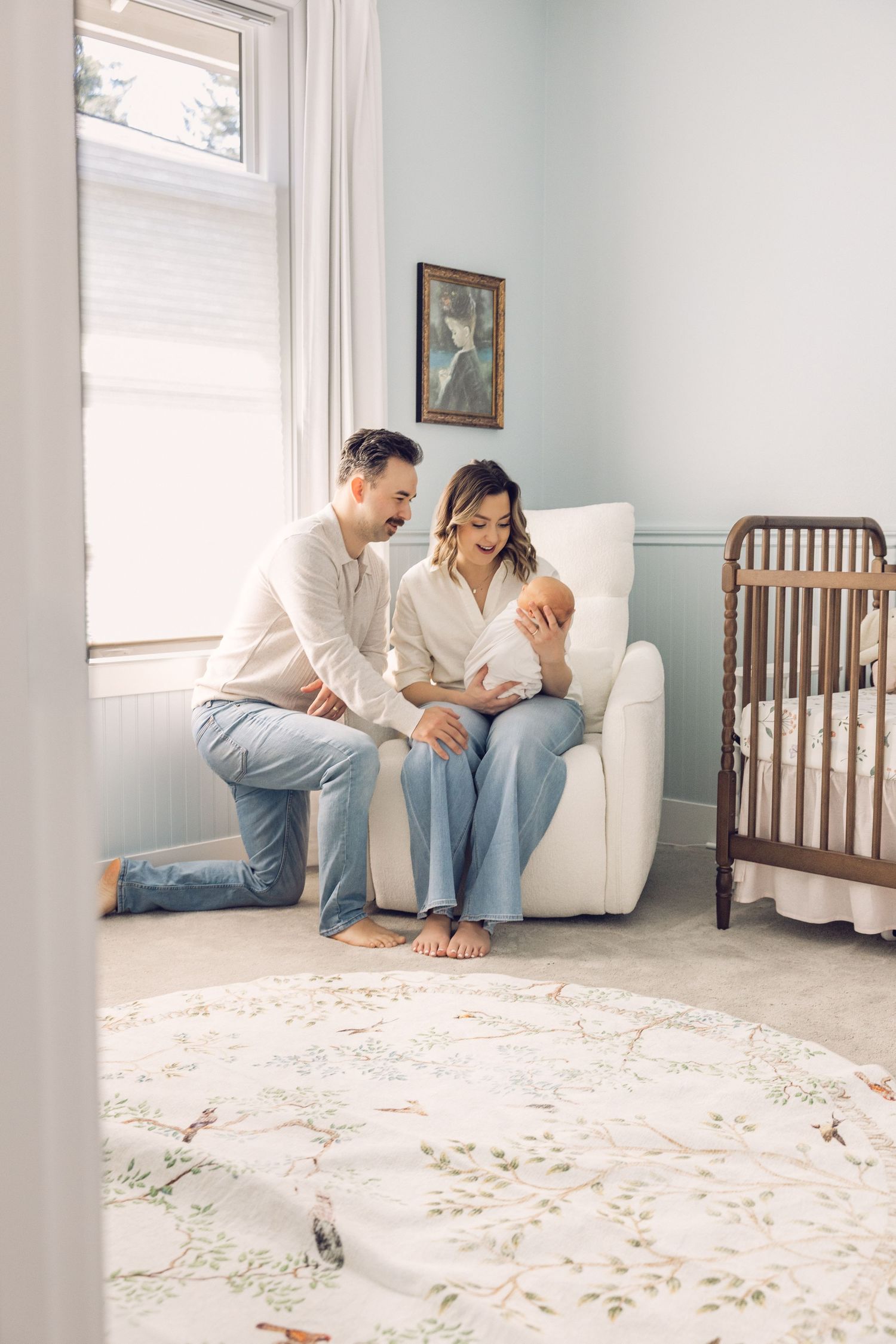 New parents admire their newborn baby in a serene nursery with soft blue walls, white glider, and wooden crib.