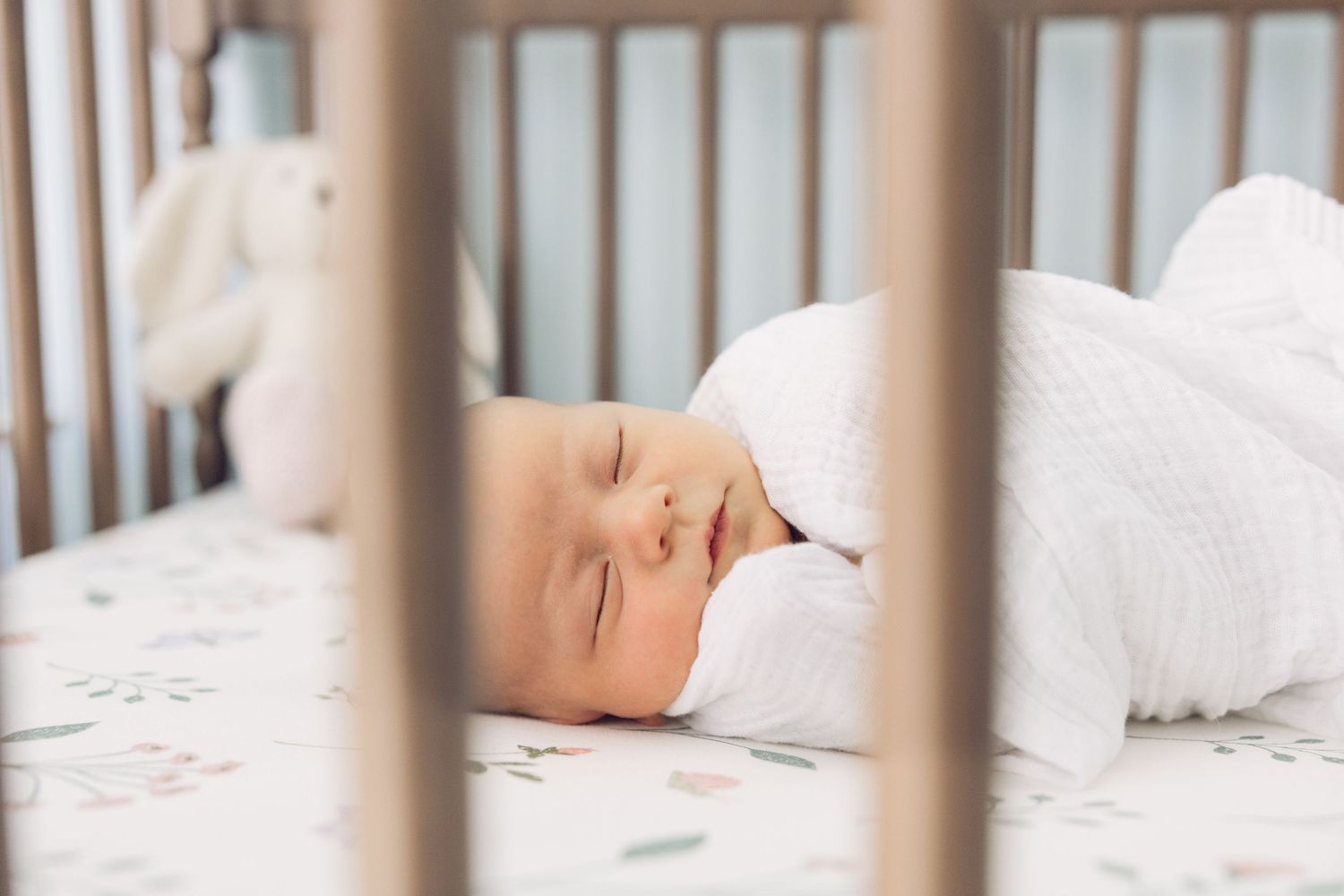 A peaceful newborn baby sleeping soundly in a white crib on floral sheets, wrapped in a soft white outfit.