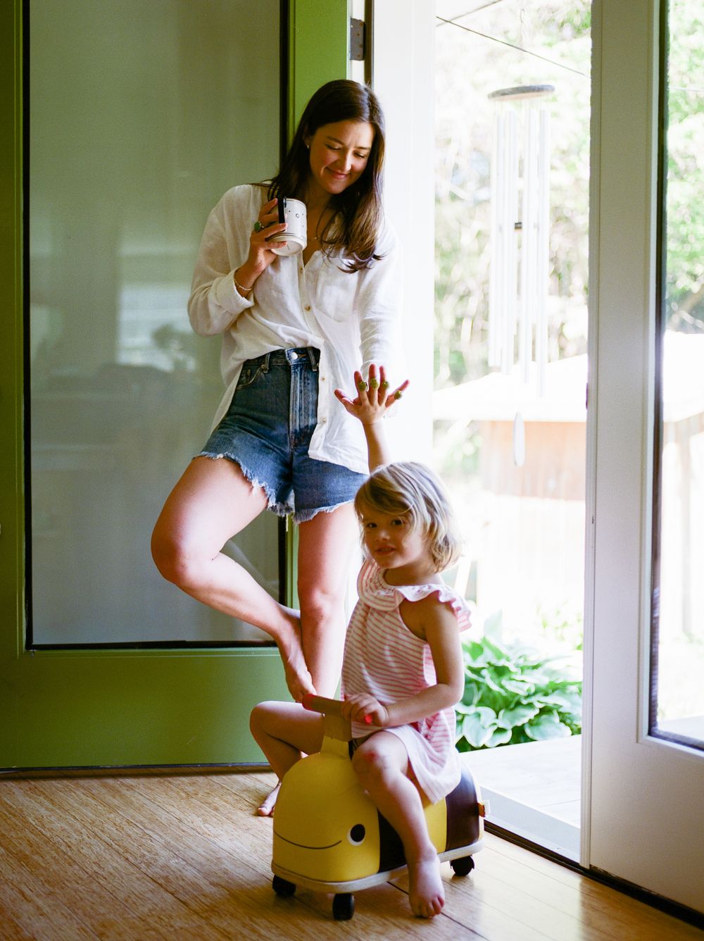 A woman in denim shorts holds a bottle while a young girl sits on a yellow ride-on toy near a glass door.