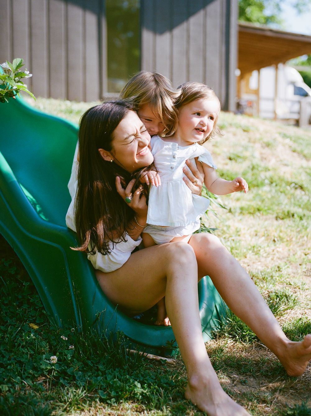 A woman sits on a green slide outdoors, hugging two young girls in white dresses on a sunny day.