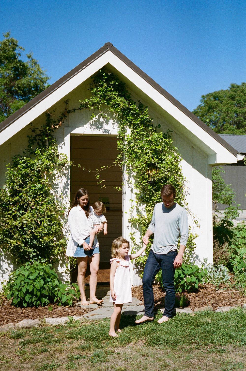 Family of four posing outside a charming white cottage covered in climbing vines on a sunny day.