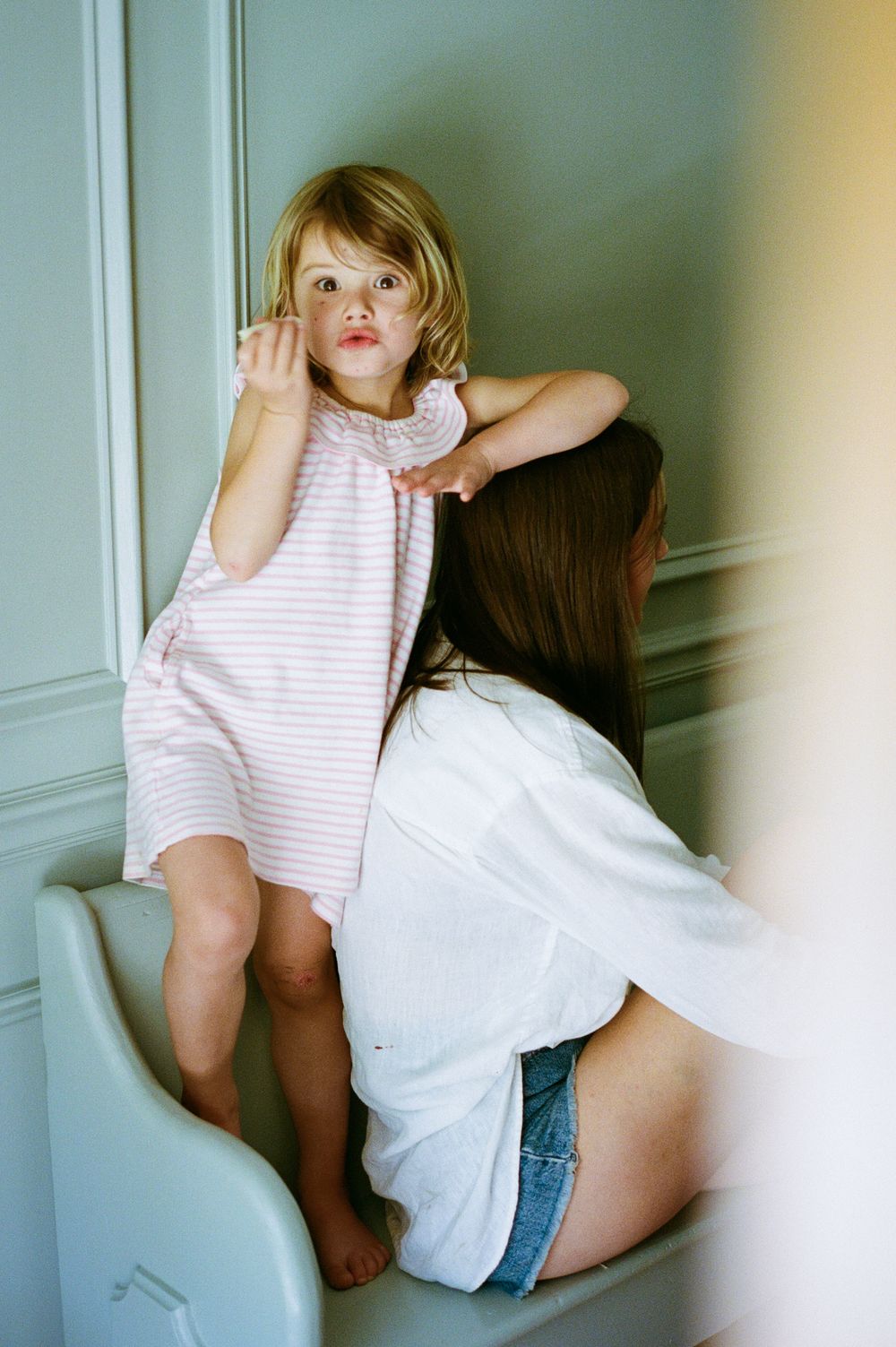 A young blonde toddler in a pink striped dress stands on a chair beside a dark-haired woman in a white shirt.
