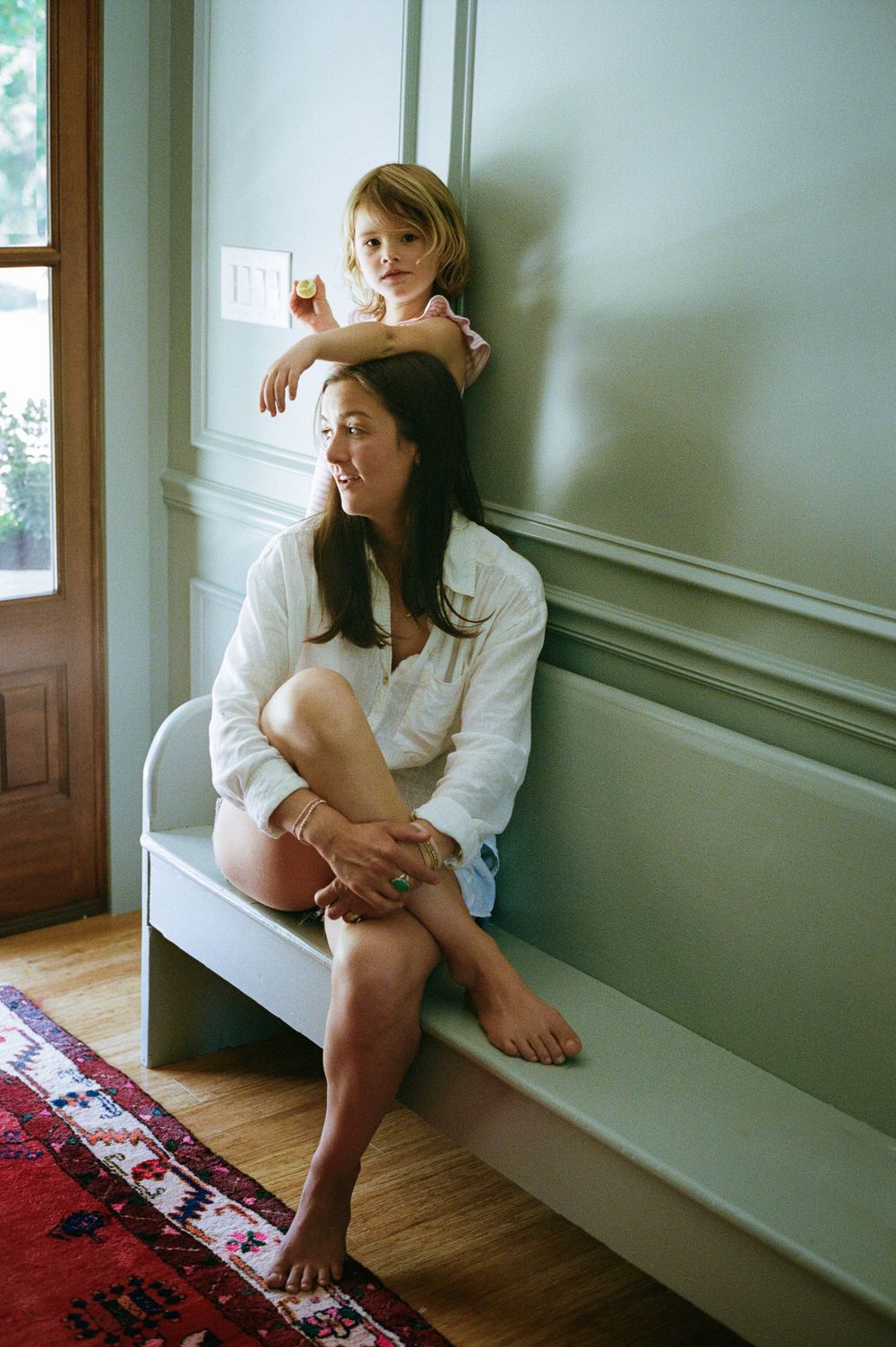 A woman sits barefoot on a bench while a young child stands behind her, both leaning against a sage green wall.