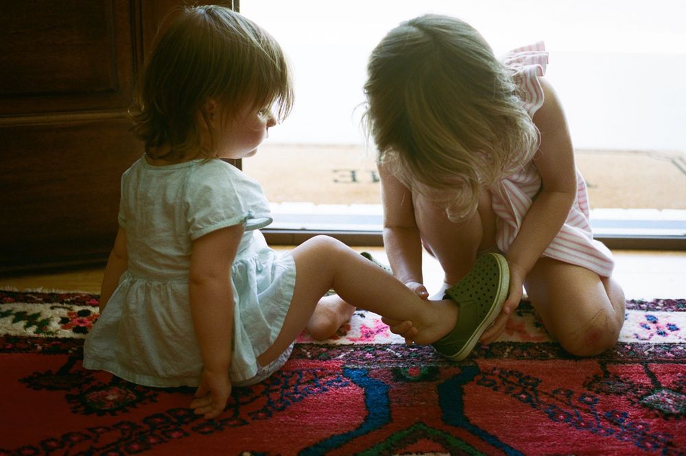 Two young girls sitting on a red patterned rug near a window, one helping the other put on a shoe.