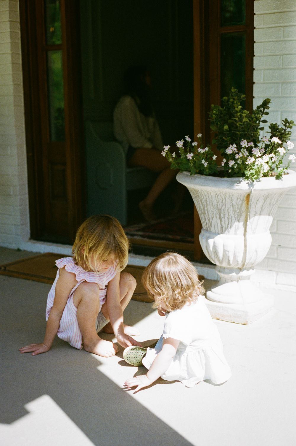 Two young toddler girls playing together on a sunlit porch beside a white urn planter with white flowers.
