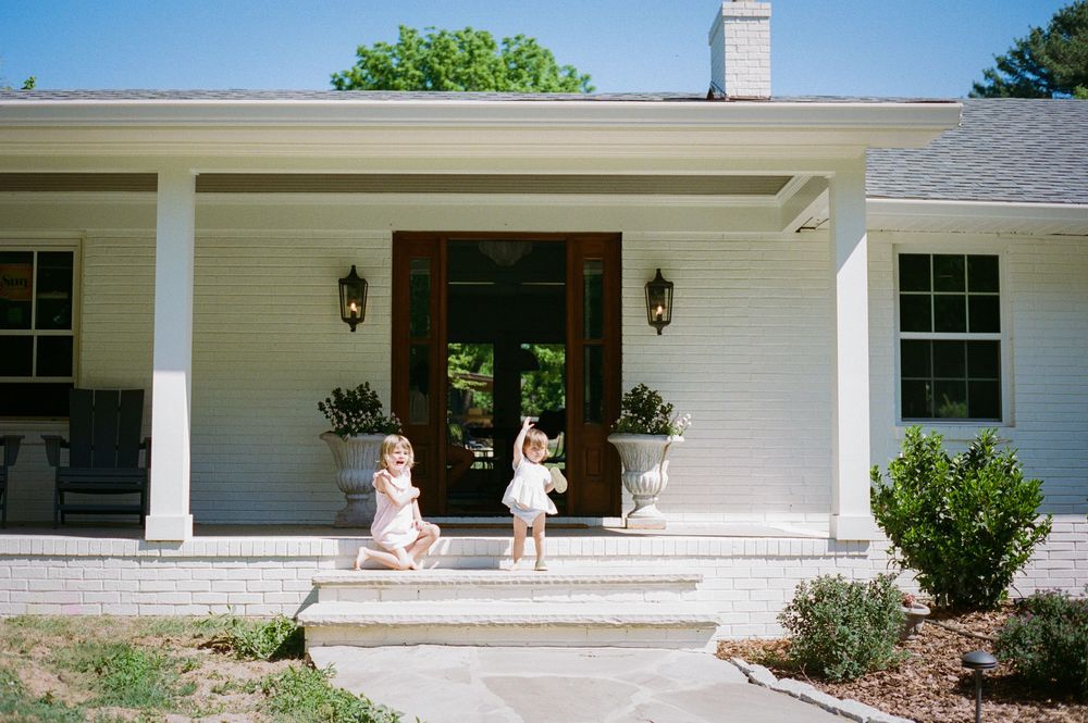 Two young children playing on the front porch steps of a white colonial-style home with a wooden double door.