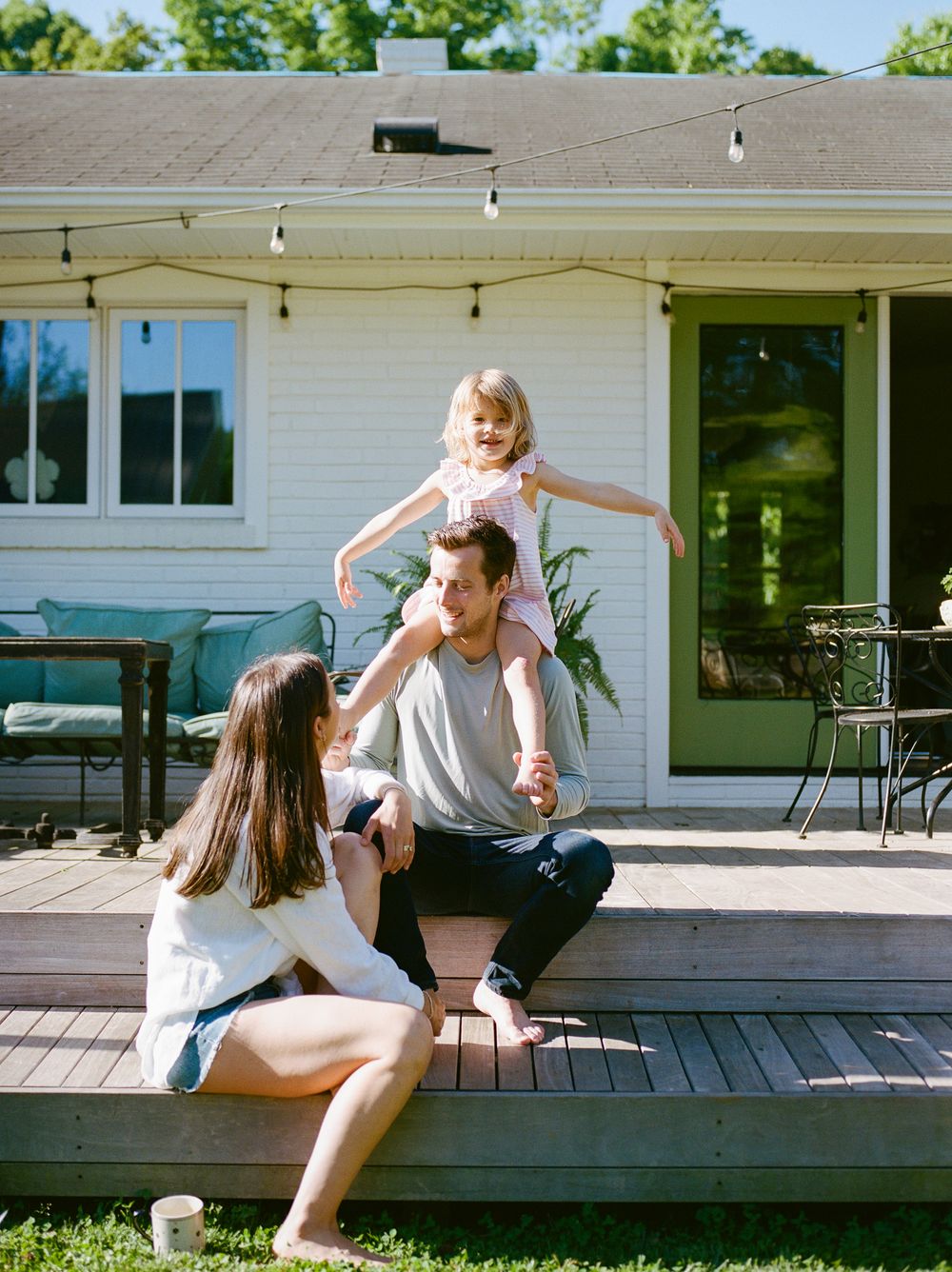 Family enjoying sunny day on backyard deck, child on father's shoulders, mother sitting nearby, cozy home exterior.